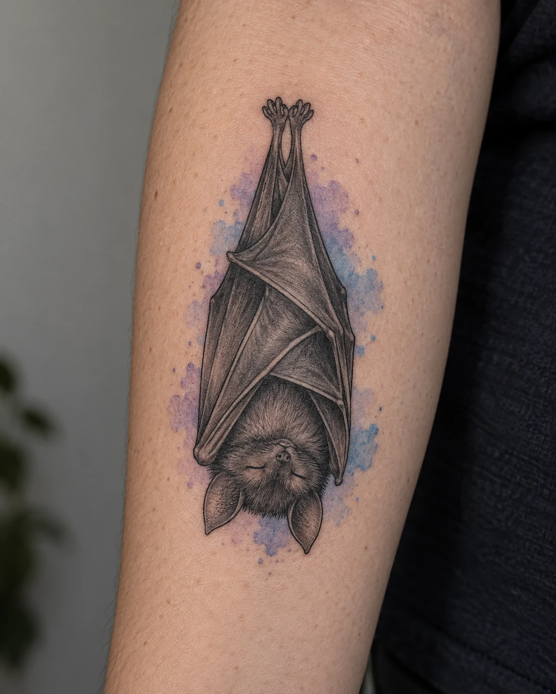 Sleeping bat tattoo with a pale lavender watercolor accent on the forearm